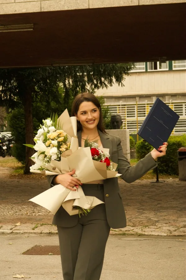 A graduate in a green trouser suit with an armful of lilies and roses, her diploma folder raised in the other hand.