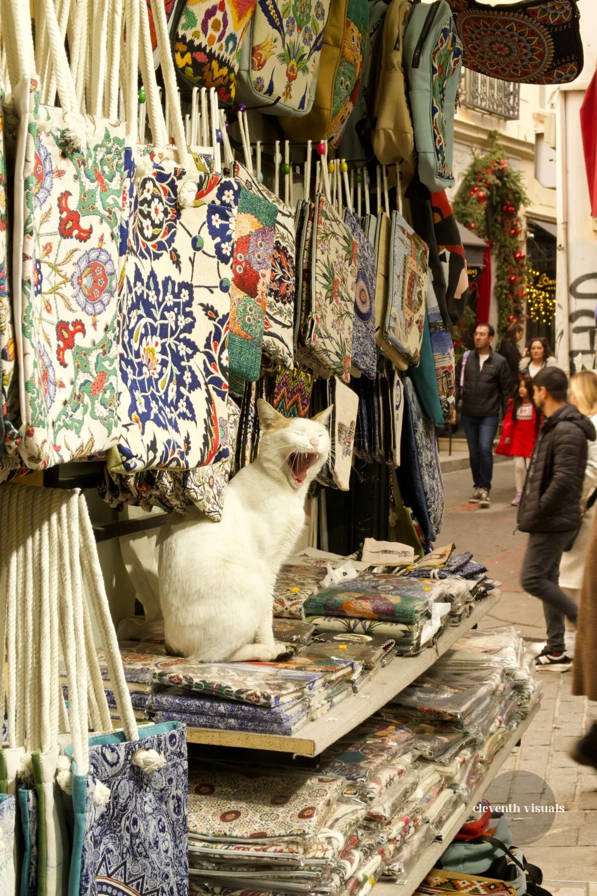 A white cat mid-yawn on a stack of printed textiles at a street stall.