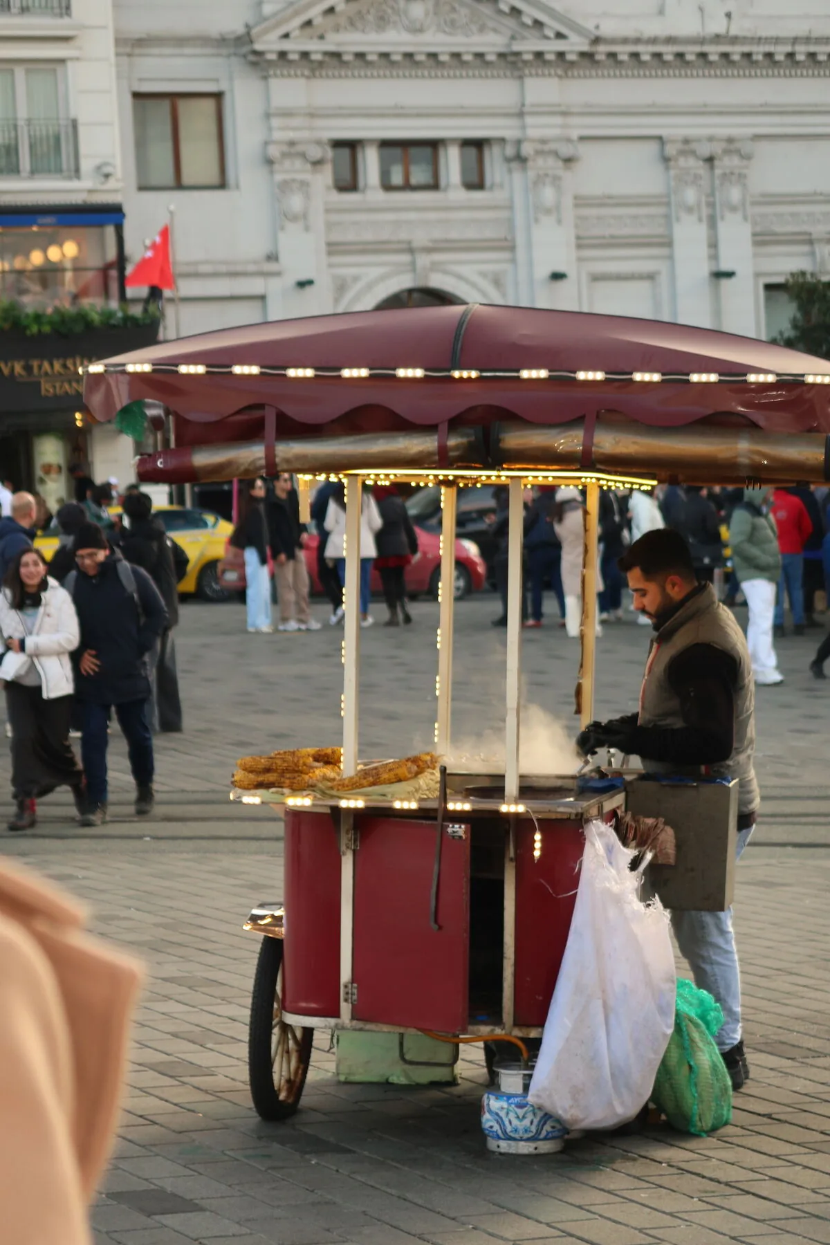 A chestnut cart with a maroon canopy and steam rising, in Taksim Square, people passing behind it.